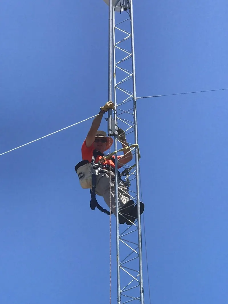 A technician in a harness climbing a lattice tower against a blue sky