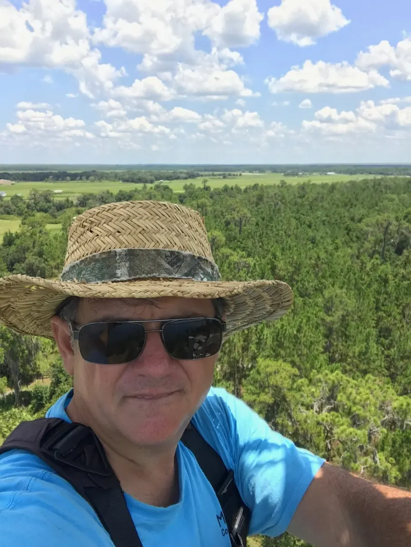 Corey Morris in a straw hat and sunglasses, high on a tower above fields and forest
