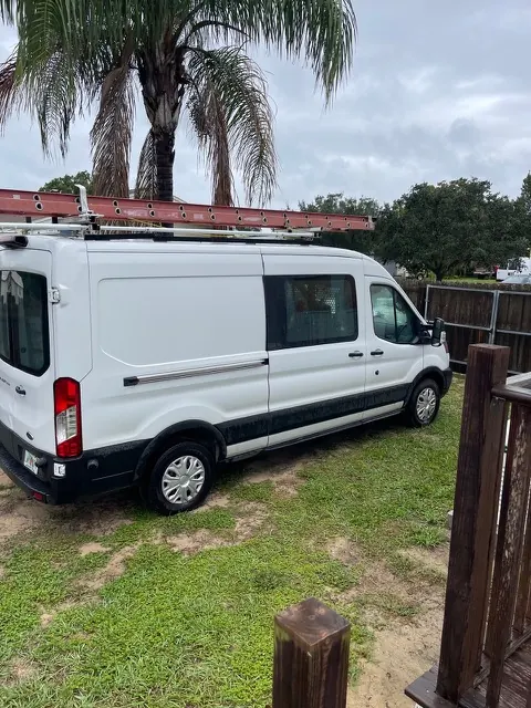 A white work van with a ladder rack parked on grass under palm trees